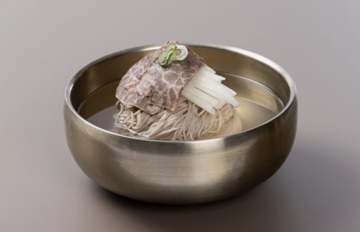 A refined brass bowl of Pyongyang naengmyeon topped with brisket slices, radish, and a scallion garnish