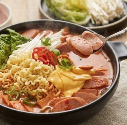 Budae-jjigae in a black pot with sausages, ramen noodles, melted cheese, chili and green onions