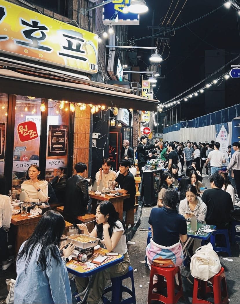 A crowded Euljiro pojangmacha alley at night with a glowing yellow beer-hall sign and diners at low tables with soju and food under warm string lights