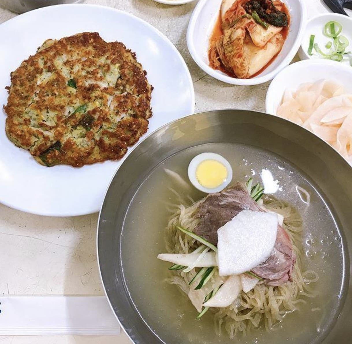 A metal bowl of Pyongyang naengmyeon in icy broth beside a golden mung-bean pancake and kimchi banchan