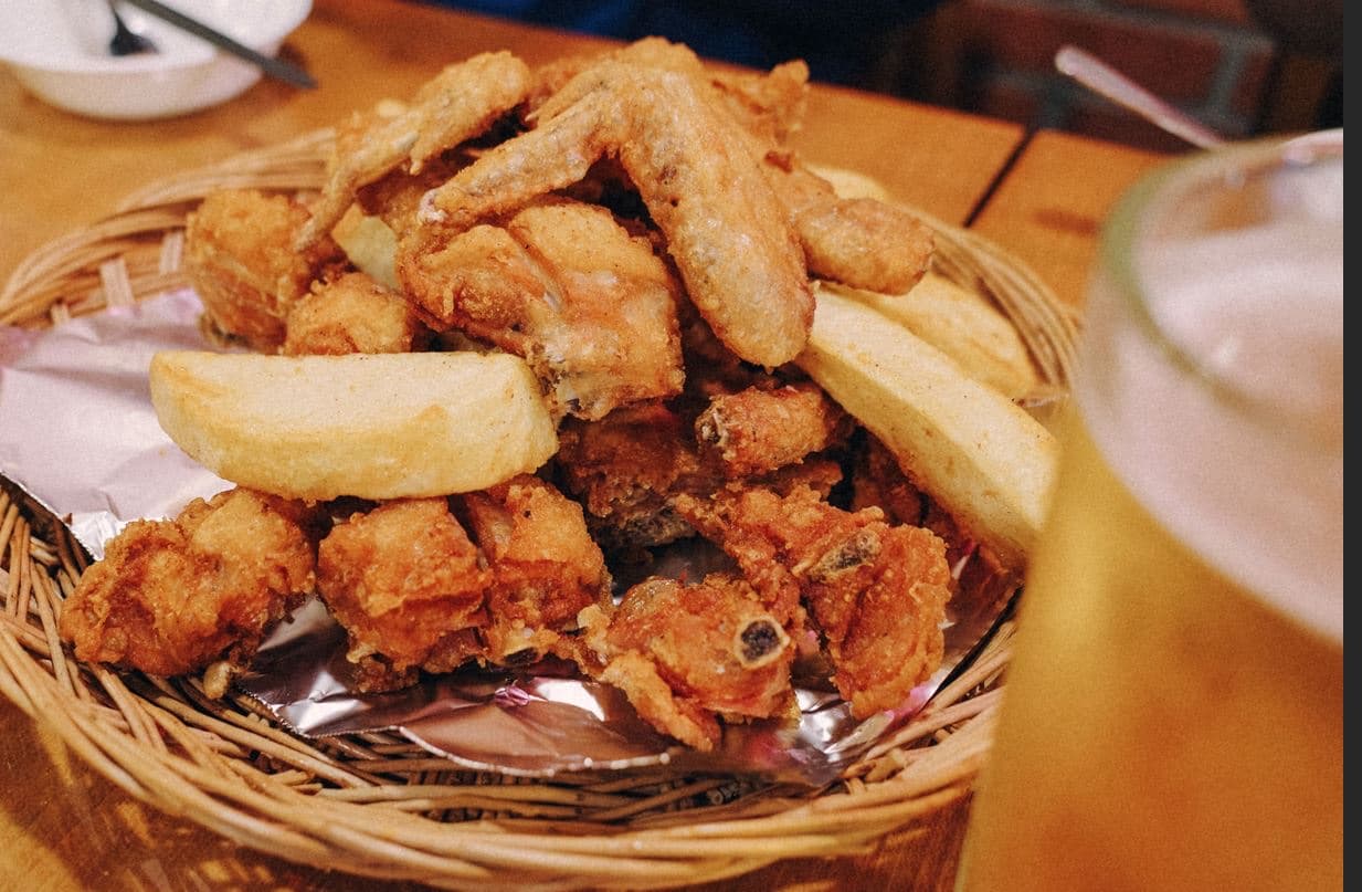 A wicker basket of crispy Korean fried chicken and thick potato wedges on foil, with a glass of draft beer alongside