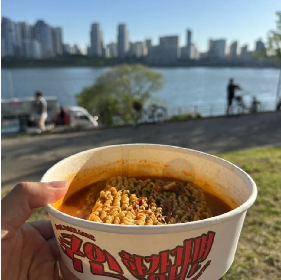 A hand holding a cup of freshly cooked Korean instant ramyeon with the Han River, riverside cyclists, and the Seoul skyline behind — the classic Hangang ramyeon experience
