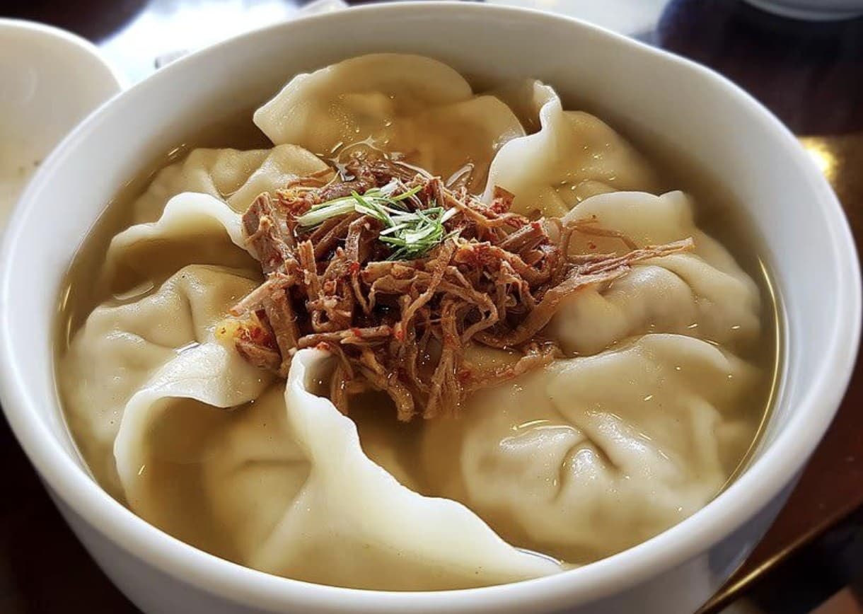 A white bowl of Jaha Sonmandu dumpling soup — plump mandu in clear broth topped with shredded seasoned beef and scallions