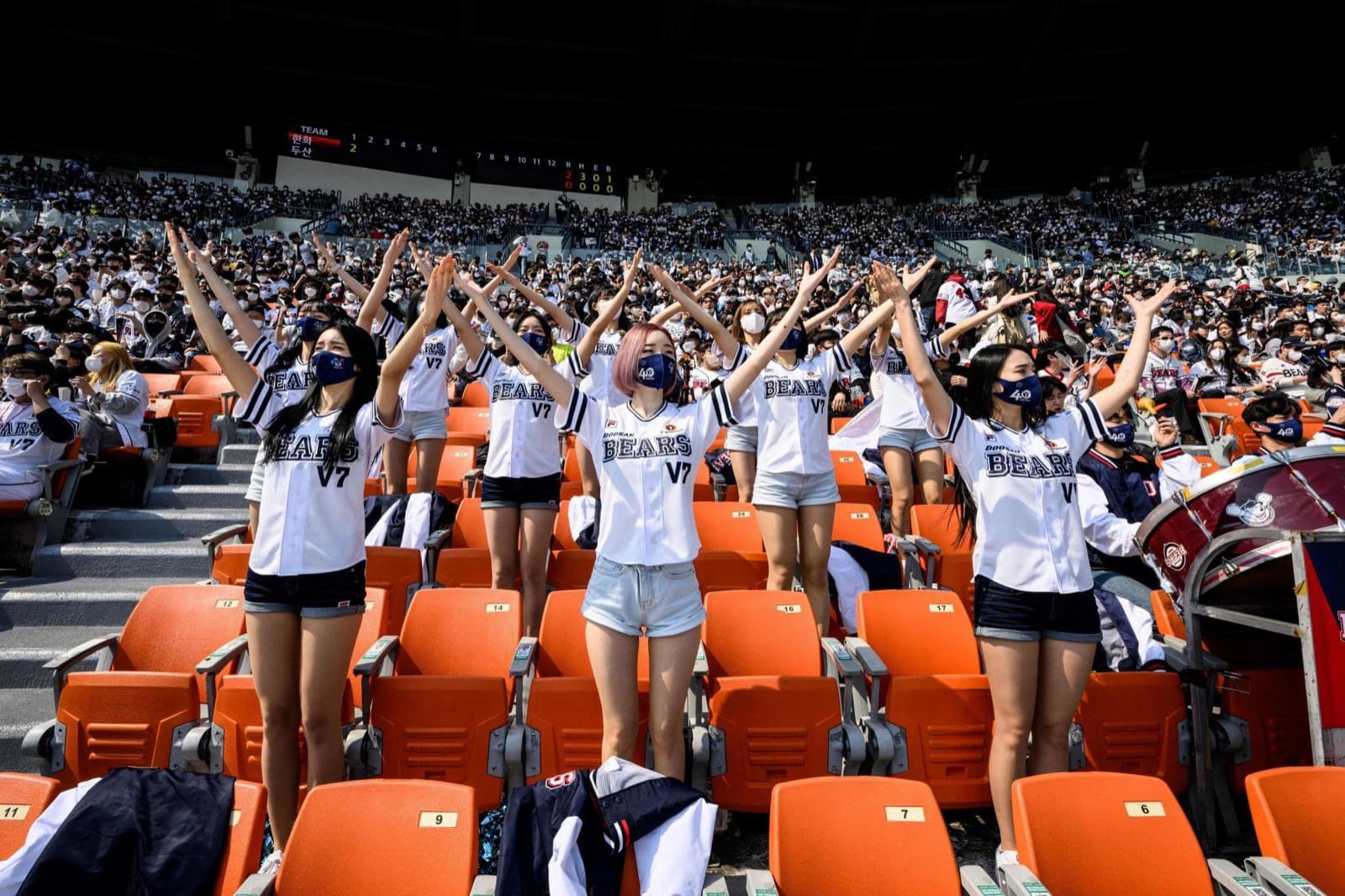 Doosan Bears fans in white BEARS V7 jerseys standing in the stands with arms raised, cheering in unison amid a packed crowd of orange seats