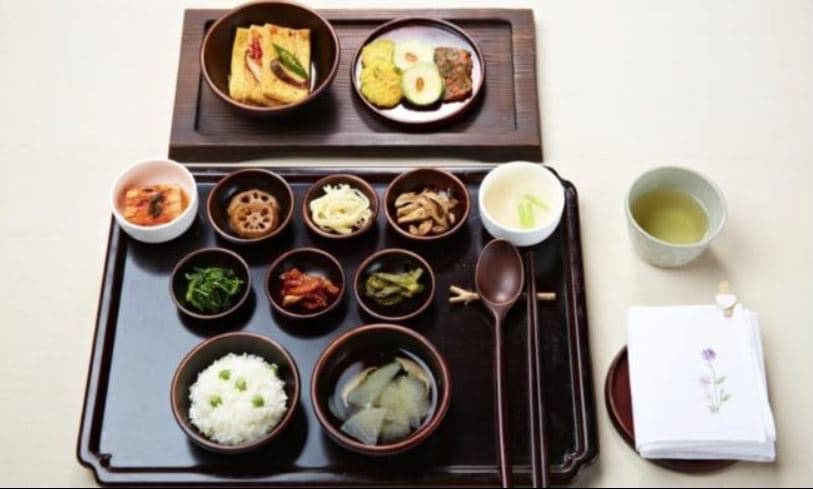 An overhead view of a Balwoo Gongyang temple meal — rice, clear soup, and many small dishes of vegetable and wild-green banchan on dark wooden trays with a cup of green tea