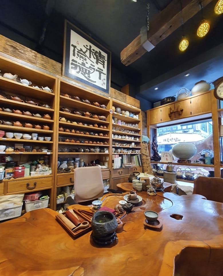 A cozy traditional tea house interior with wooden shelves full of teapots and cups, a large natural-wood tea table, and warm lighting