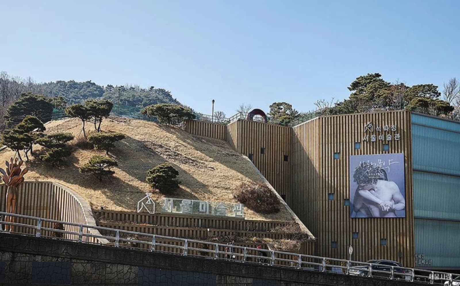 The Seokpajeong Seoul Museum building with a gold vertical-slat facade set into a grassy hillside, with an exhibition banner on the side