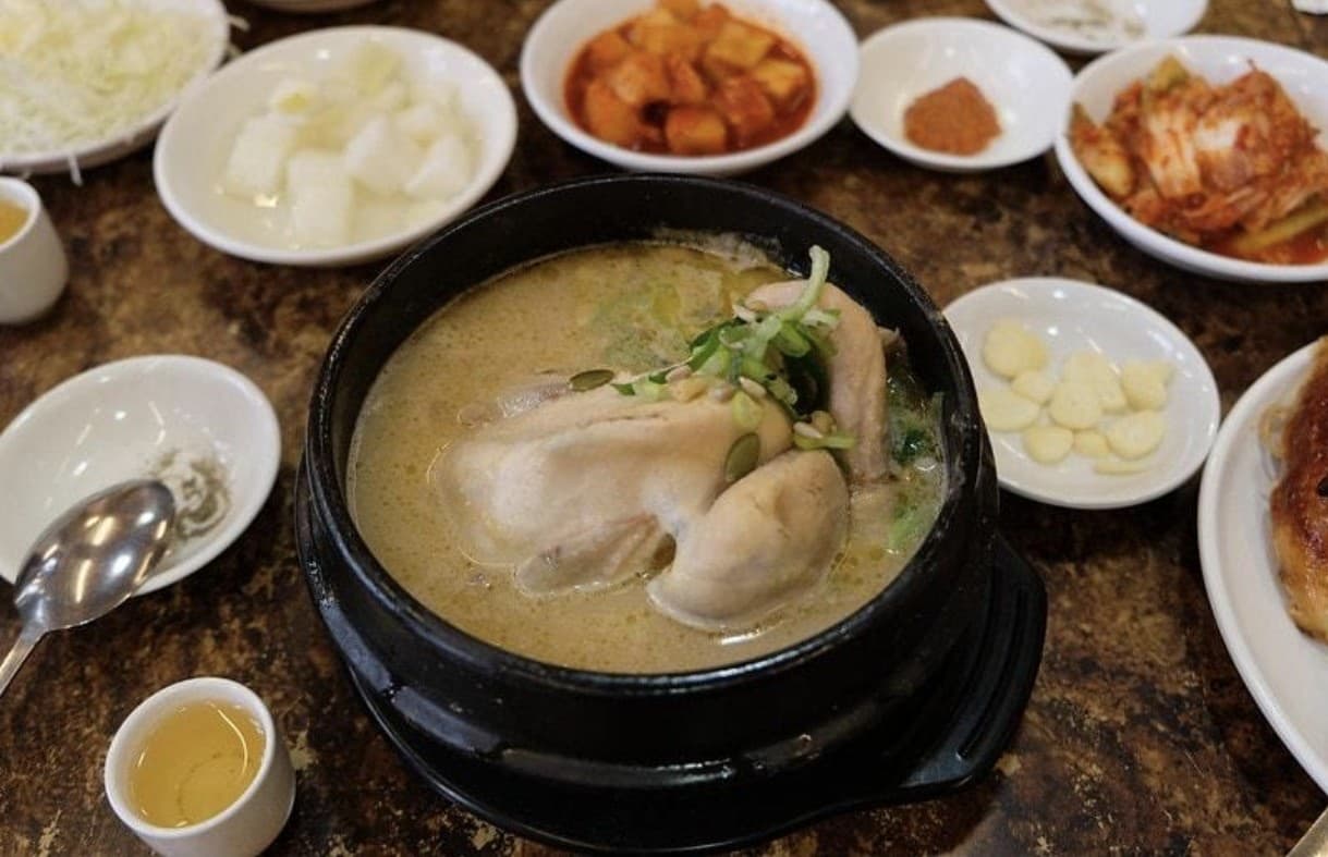 A stone pot of Tosokchon samgyetang — whole chicken in milky ginseng broth topped with scallions, surrounded by banchan side dishes