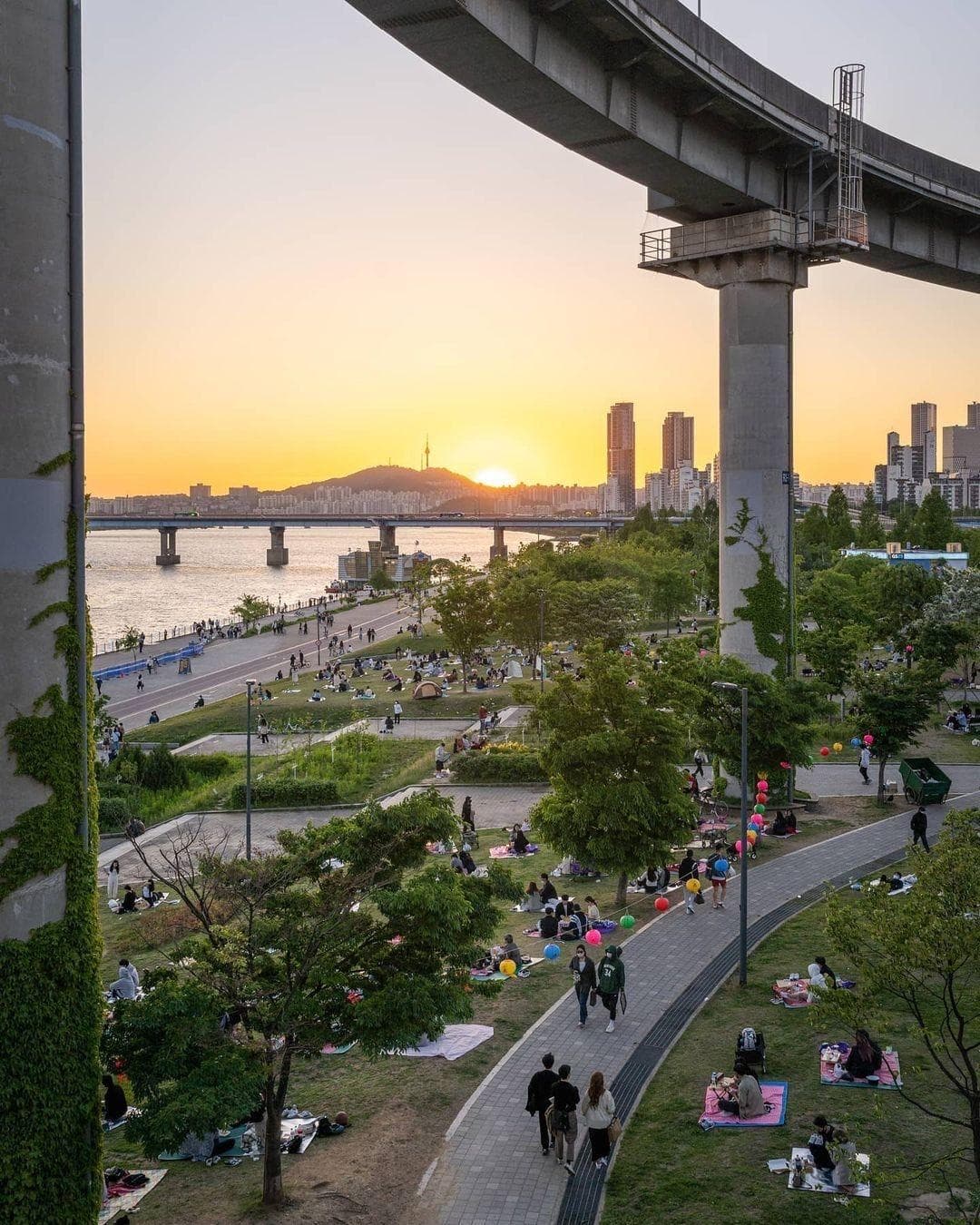 Golden-hour view of Ttukseom Hangang Park with crowds picnicking on riverside lawns, colorful lanterns, the elevated monorail overhead, and the Seoul skyline with N Seoul Tower at sunset