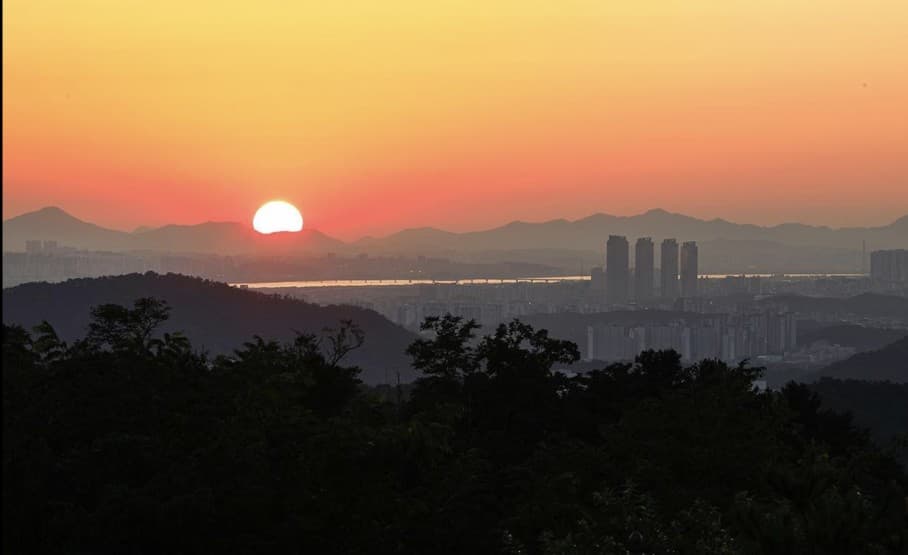The sun setting behind layered mountain ridges above the hazy Seoul cityscape and river